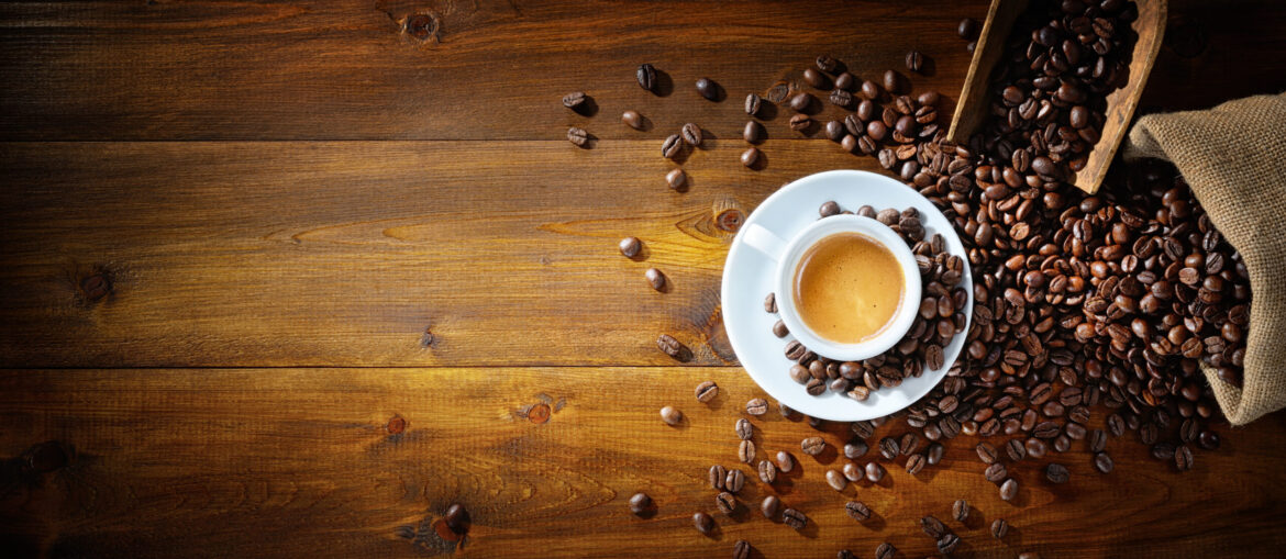 White cup of espresso on a saucer surrounded by scattered coffee beans on a rustic wooden table, with a burlap bag spilling beans on the right.