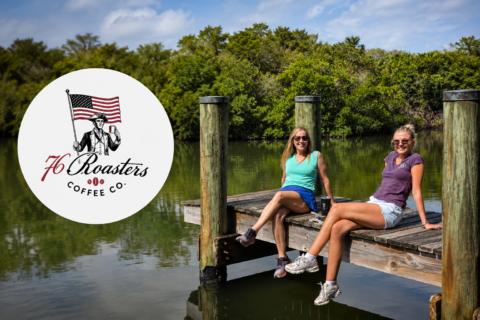 Two women sit on a wooden dock by a calm lake, smiling in sunglasses; a large white circle logo for 76 Roasters Coffee Co. sits on the left edge of the frame.